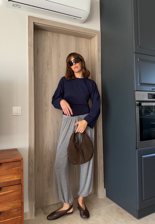 Femme portant des lunettes de soleil noires, un haut à manches longues bleu marine, un pantalon gris ample, des chaussures tressées marron et tenant un sac à main marron, debout près d'une porte en bois.