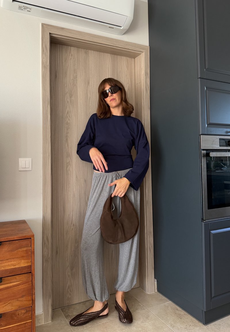 Femme portant des lunettes de soleil noires, un haut à manches longues bleu marine, un pantalon gris ample, des chaussures tressées marron et tenant un sac à main marron, debout près d'une porte en bois.