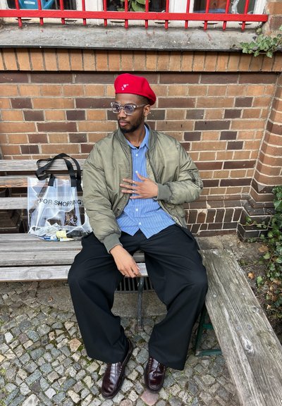 Hombre con un boina roja, chaqueta bomber verde oliva, camisa de rayas azul y pantalones anchos negros, sentado en un banco de madera con una bolsa transparente.