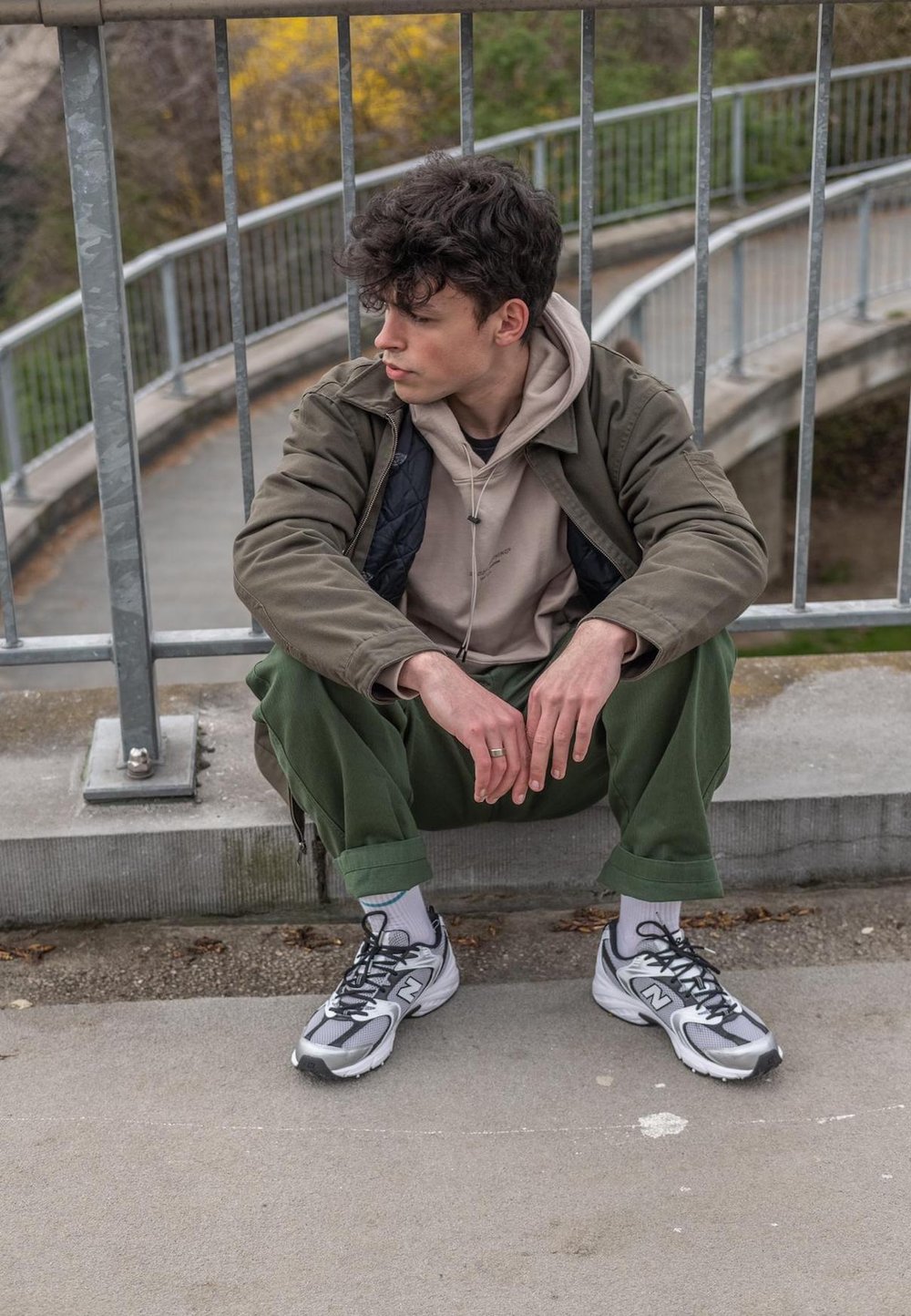 Pohodlný outfit pozostávajúci z béžovej mikiny pod olivovo zelenou bundou, zelených nohavíc a šedých teniskách New Balance, sediac na betónovom povrchu.