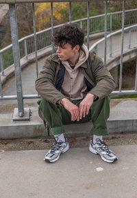 Pohodlný outfit pozostávajúci z béžovej mikiny pod olivovo zelenou bundou, zelených nohavíc a šedých teniskách New Balance, sediac na betónovom povrchu.