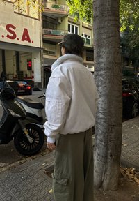 Valge ülisuure bomber-tüüpi jakk fliisvoodriga kraega, kombineeritud oliivroheliste taskupükstega. Stseenil on puud ja pargitud sõidukid.