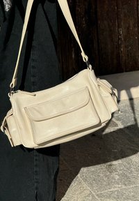 Beidzs ādas neliela crossbody soma ar rāvējslēdzēja aizdari, priekšējo kabatu un sānu nodalījumu, kurai ir plakana plecu siksna un gluda tekstūra.