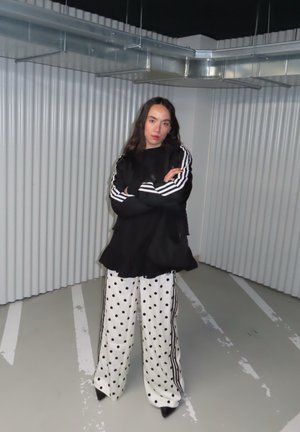 Mujer de cabello largo y oscuro de pie con los brazos cruzados, vistiendo una blusa negra con rayas blancas y pantalones anchos blancos con lunares en un entorno industrial.