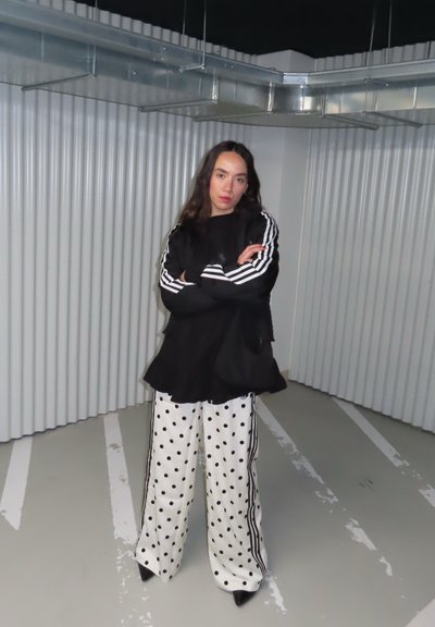 Mujer de cabello largo y oscuro de pie con los brazos cruzados, vistiendo una blusa negra con rayas blancas y pantalones anchos blancos con lunares en un entorno industrial.