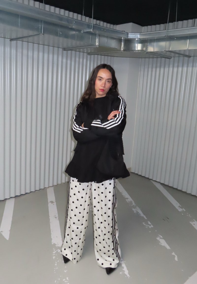 Femeie cu păr lung și întunecat, stând cu brațele încrucișate, purtând un top negru cu dungi albe și pantaloni largi albi cu buline, într-un cadru industrial.