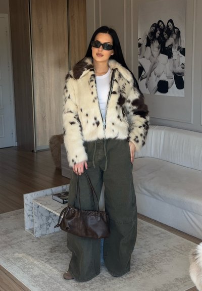 Chaqueta de piel sintética con patrón de manchas marrones, cierre de cremallera y mangas anchas. Combinada con pantalones olive verdes sueltos y un pequeño bolso marrón.