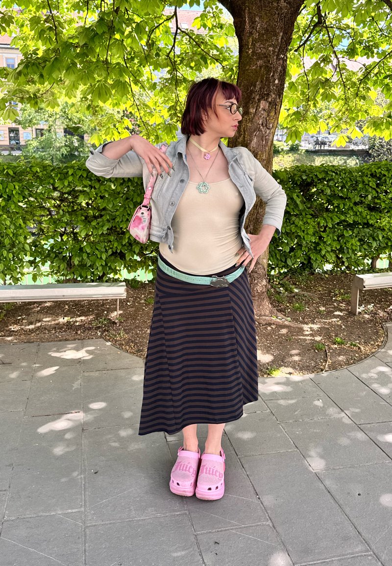 Personne aux cheveux courts, haut beige, jupe rayée, chaussures roses et sac, debout sous un arbre feuillu sur un chemin pavé avec des bancs à proximité.