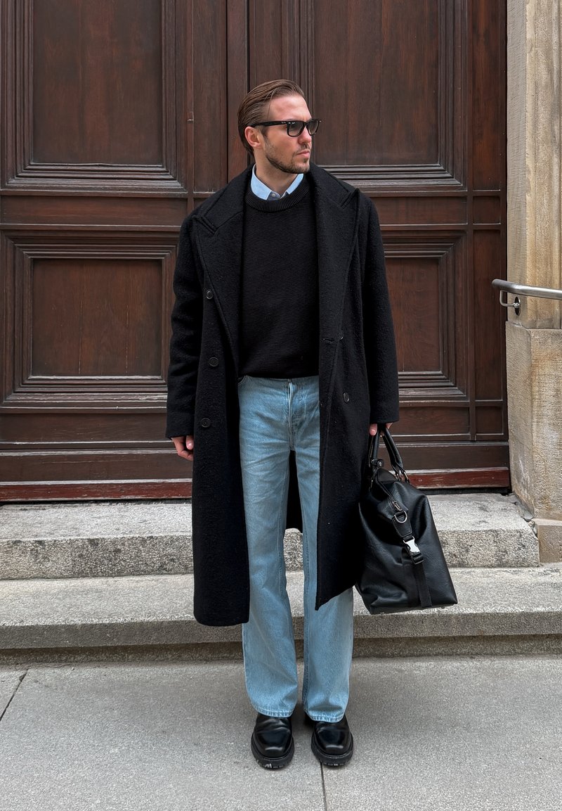 Homme portant un manteau noir, un pull noir, un jean bleu clair et des lunettes de soleil, tenant un sac en cuir noir, debout sur des marches en pierre devant une porte en bois.