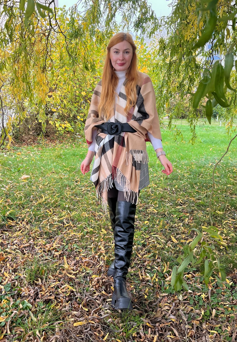 Poncho à carreaux beige et noir avec un ourlet à franges, cintré à la taille par une ceinture noire ; associé à des bottes noires montantes sur un sol herbeux.