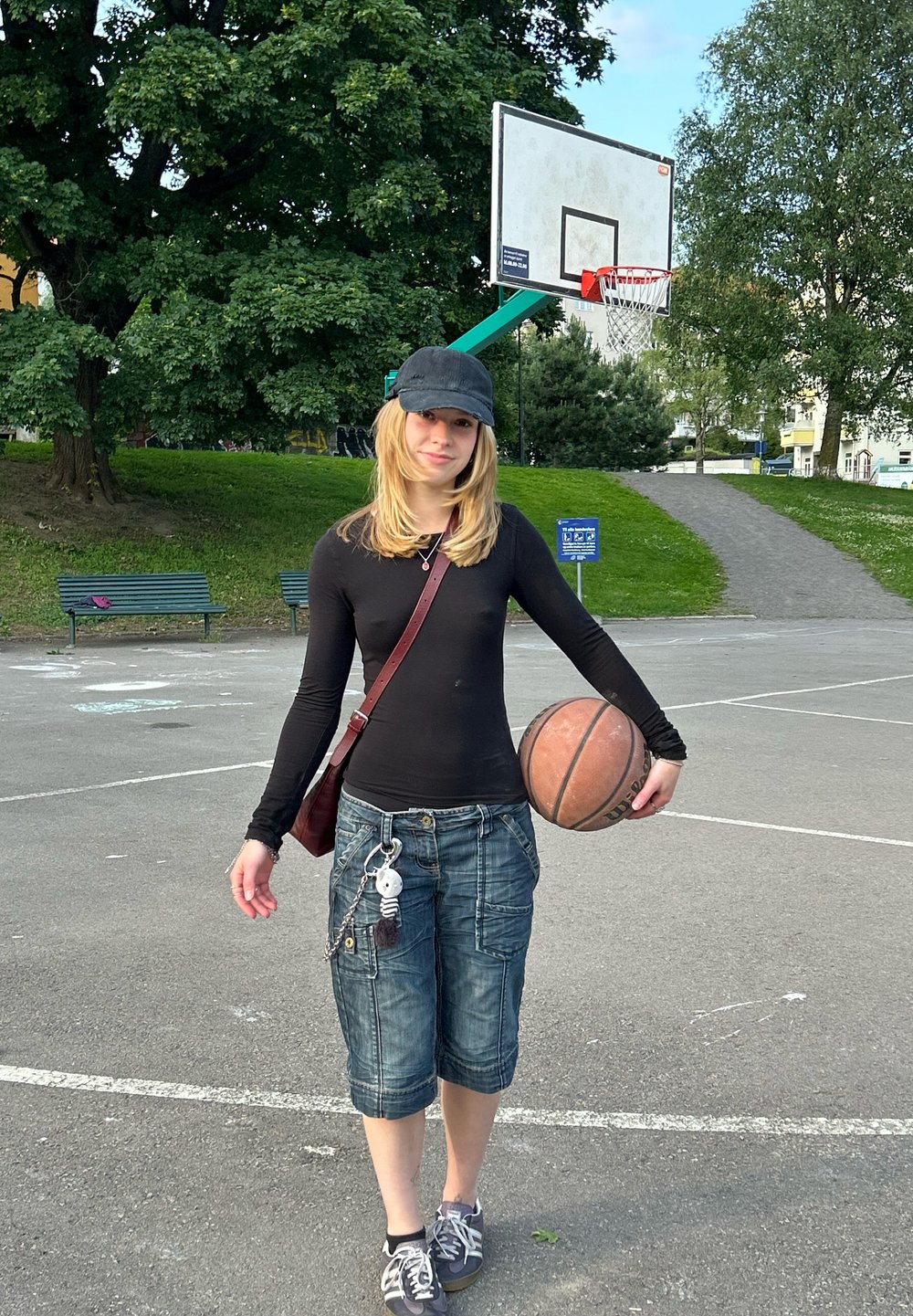 Basketbollspelare som håller en brun boll, iklädd en svart långärmad tröja, denim capribyxor och svarta sneakers, med en svart keps.