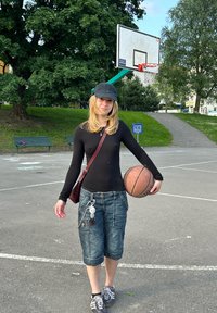 Basketbollspelare som håller en brun boll, iklädd en svart långärmad tröja, denim capribyxor och svarta sneakers, med en svart keps.