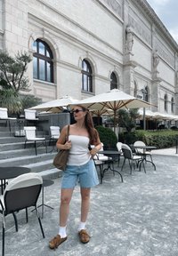 Gaišs brūns plecu soma, balta T-kreklu, denima šorti un brūni mokasīnu stila apavi. Apkārt galdi un lietussargi.