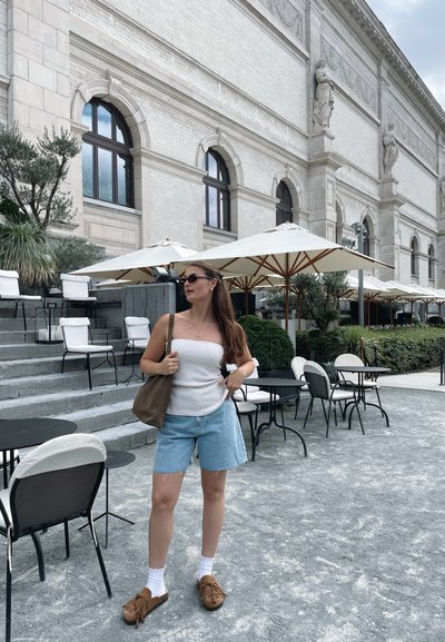 Bolso de hombro marrón ligero, camiseta sin mangas blanca, pantalones cortos de mezclilla y zapatos estilo mocasín marrones. Rodeado de mesas y sombrillas.