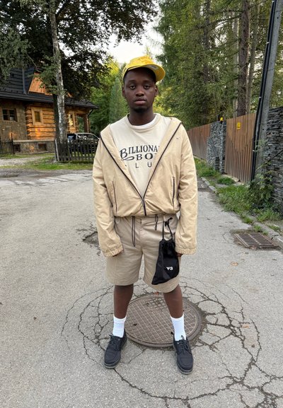 Chaqueta bomber beige con capucha, camiseta blanca, pantalones cortos caqui, bolsa negra con cordón, calcetines blancos y zapatos negros. Entorno al aire libre.