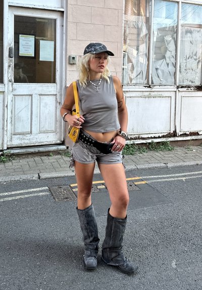 Top de tirantes gris corto, pantalones cortos grises con flecos laterales, riñonera negra con tachuelas, bolso amarillo, botas gris hasta la rodilla y una gorra estampada.