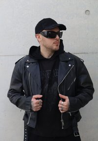 Fekete bőr motoros dzseki ezüst cipzárakkal, sötét kapucnis pulóver fölött viselve. Kiegészítők közé tartozik napszemüveg és több gyűrű.