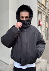 Chaqueta acolchada negra con un cuello alto y una capucha grande. Presenta un corte holgado, textura suave y cremalleras en el dobladillo.