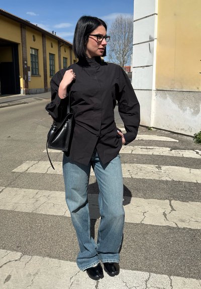 Mujer con cabello negro y gafas está parada en un paso de peatones, lleva una camiseta negra, vaqueros azules, zapatos negros con dedo dividido y sostiene un bolso negro.