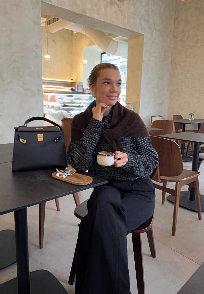 Ung kvinne på en kafé som holder en kopp kaffe, med svart håndveske og øretelefoner på bordet, sitter på en trestol med lysmalte vegger.