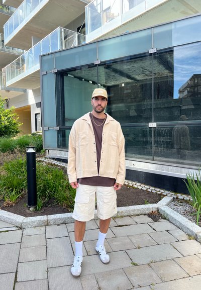 Chaqueta de pana beige sobre una camiseta marrón, pantalones cortos cargo blancos, zapatillas blancas y una gorra beige. De pie en un entorno urbano al aire libre.