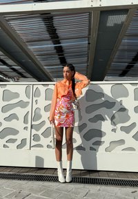 Giacca cropped leggera arancione sopra un vestito mini a fantasia floreale colorato, con stivali bianchi fino alla coscia e mentre tiene una piccola borsa bianca.