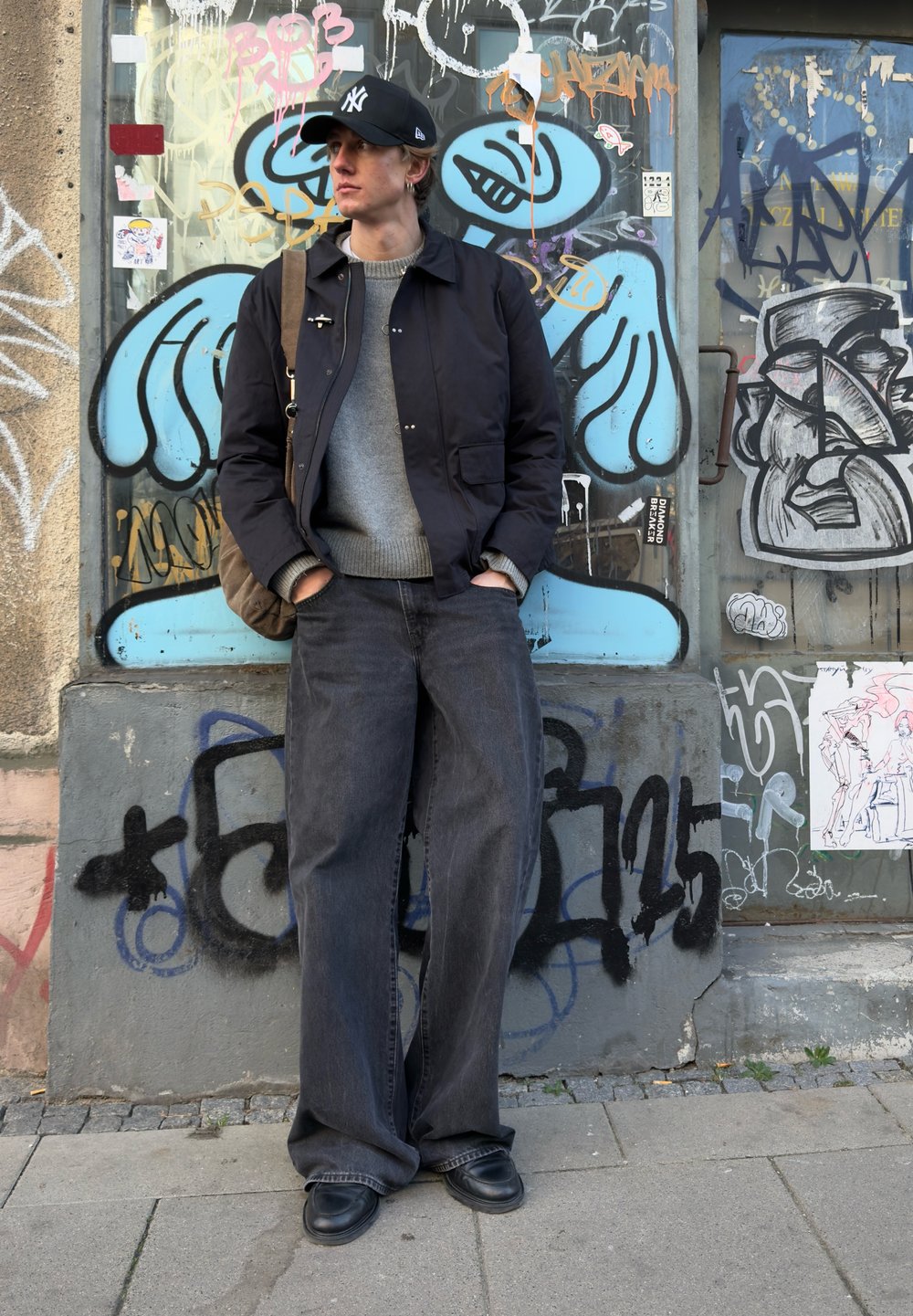 Giacca bomber nera sopra un maglione grigio, abbinata a jeans in denim scuro a gamba larga e scarpe nere. In piedi davanti a un muro con graffiti colorati.