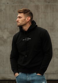 Black hoodie with drawstrings, featuring white text on the chest. Worn over light blue jeans, the fabric appears soft and textured.