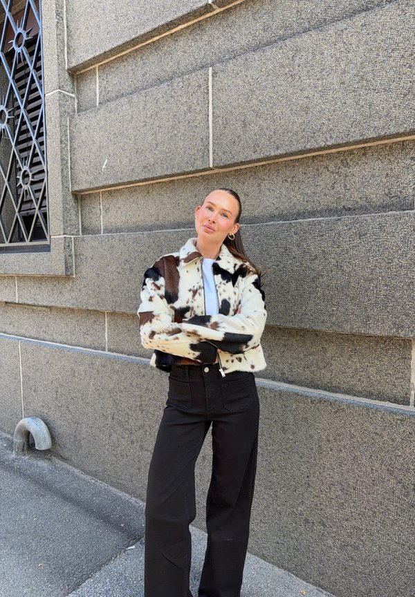 Mujer de pie con los brazos cruzados, vestida con una chaqueta negra y blanca con manchas y pantalones negros contra una pared de piedra gris texturizada.