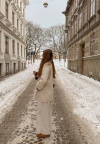 Άτομο με μπεζ παλτό και παντελόνι στέκεται σε δρομάκι με πλακόστρωτο καλυμμένο με χιόνι ανάμεσα σε ιστορικά κτίρια με γυμνά δέντρα το χειμώνα.