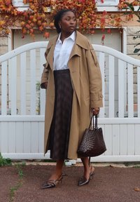 Beżowy trencz na białej koszuli, brązowa spódnica w kratę. Dopełnieniem stroju jest brązowa pleciona torebka i szpilki z zaostrzonym noskiem. W tle biały drewniany płot.