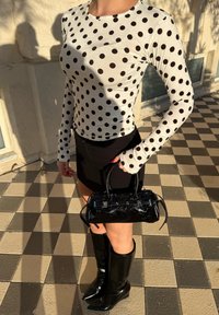 Femme portant une chemise blanche à manches longues avec des pois noirs, une mini-jupe noire, des bottes noires montantes jusqu'aux genoux, et tenant un petit sac à main en cuir verni noir.