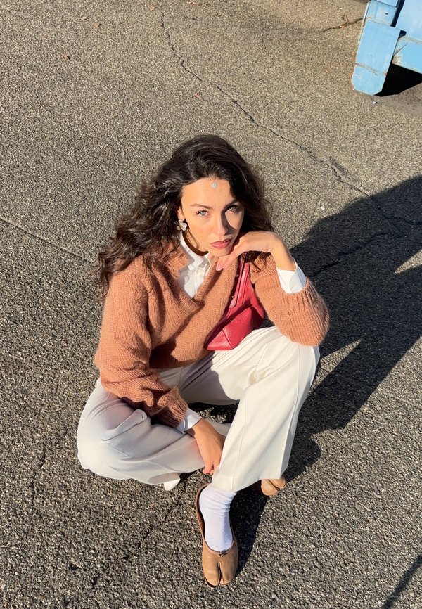 Maglione a maglia marrone, camicia bianca, pantaloni bianchi larghi e ballerine marrone chiaro. Tiene una piccola borsa rossa, seduta su una superficie grigia testurizzata.