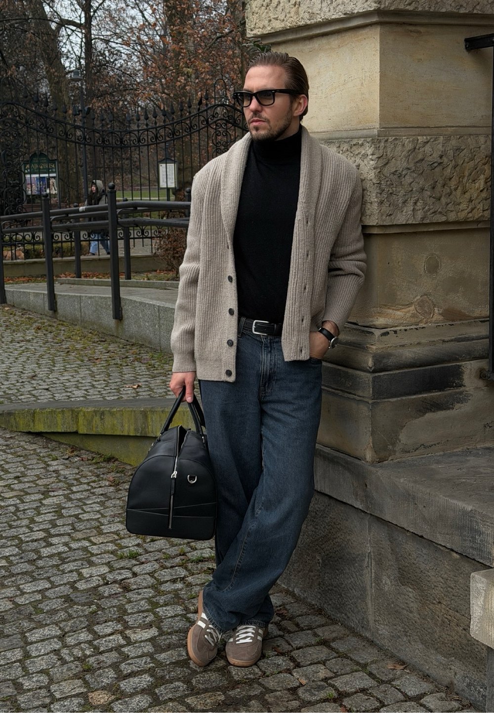 Cardigan gris clair côtelé porté sur un col roulé noir, associé à un jean bleu foncé et des baskets beige. Tenant un sac en cuir noir.