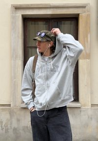 Jauns cilvēks ar pelēku kapuci, kamuflāžas cepuri, saulesbrillēm uz cepures, austiņām un plecu soma stāv pie loga uz bēša sienas.