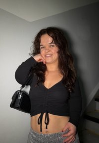 Femme aux longs cheveux foncés portant un crop top noir et une jupe à paillettes argentées, tenant un sac à main noir sur l'épaule, souriant en intérieur.