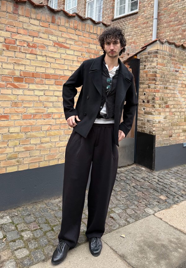 Jaunas vyras su garbanotais plaukais stovi prie plytų sienos, dėvėdamas juodus plačius kelnes, juodą paltą, kelias sluoksniuotas palaidines ir juodus batus.