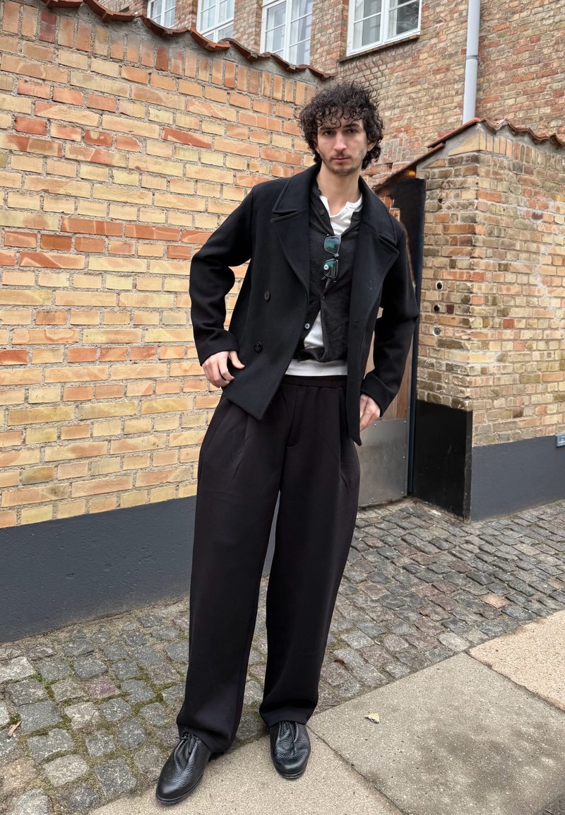 Jeune homme aux cheveux bouclés se tenant contre un mur en briques, portant un pantalon noir à jambes larges, un manteau noir, plusieurs couches de hauts et des chaussures noires.