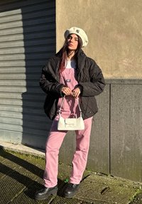 Czarna pikowana kurtka, biały beret z logo, różowe ogrodniczki, biała torebka z kokardą, czarne masywne buty, stojąca przy ścianie.