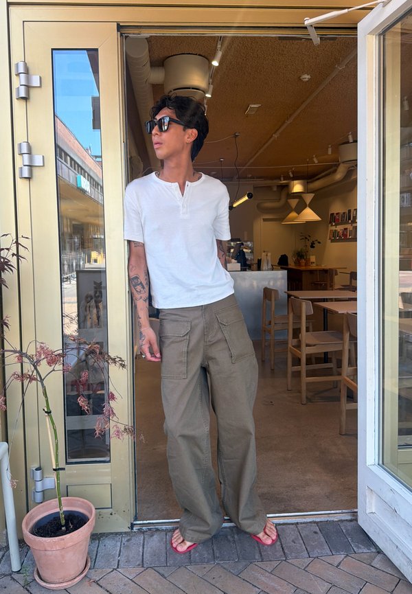 Junge Person mit Sonnenbrille, weißem Hemd und weiten Hosen steht in der offenen Tür eines Cafés mit Holztischen und -stühlen im Inneren.