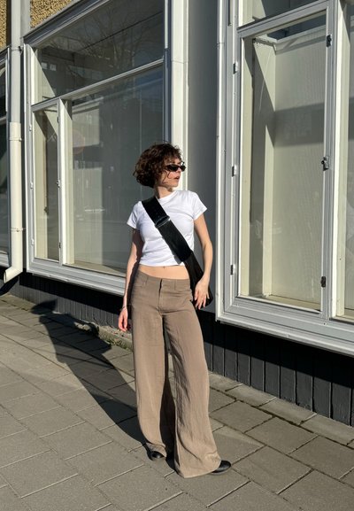 Camiseta blanca ajustada, pantalones de pierna ancha beige, bolso bandolera negro con hebilla plateada, gafas de sol, de pie en una acera near grandes ventanales.