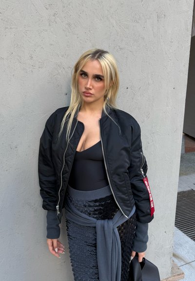 Chaqueta bomber negra con cremalleras plateadas, llevada sobre un body negro; falda texturizada y en capas de color gris oscuro, sujeta con un abrigo a juego.