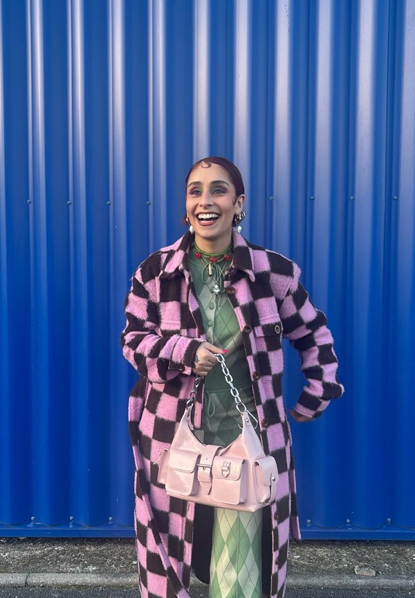 Ung kvinde smilende, iført en pink og sort ternet frakke, grønt mønstret outfit, holder en lyserød håndtaske og står foran en blå rillede væg.