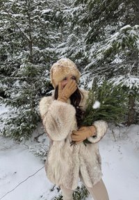 Femme en manteau de fourrure, chapeau et gants tenant des branches de conifères, se couvrant la bouche, debout dans une forêt enneigée avec de la neige sur les arbres et le sol.