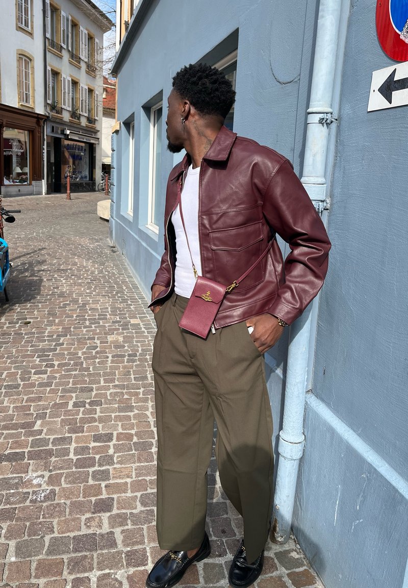 Veste en cuir bordeaux avec deux poches avant, associée à un pantalon large vert olive et des mocassins noirs. Petit sac rouge porté en bandoulière.