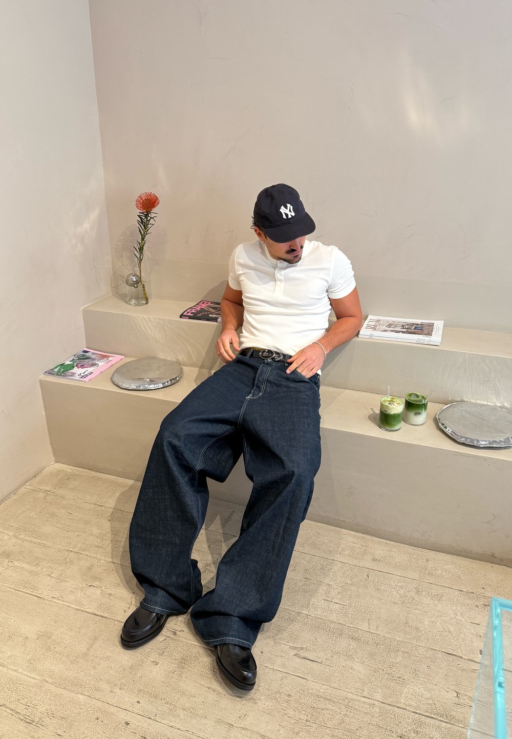 Vidbenede denimjeans i mørkeblå med en glat tekstur, kombineret med en tætsiddende hvid polo. En sort kasket og blanke sko fuldender looket.