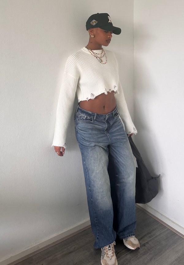 White ribbed crop sweater with frayed hem, paired with light-wash baggy jeans and beige sneakers. Accessorized with layered necklaces and a black cap.
