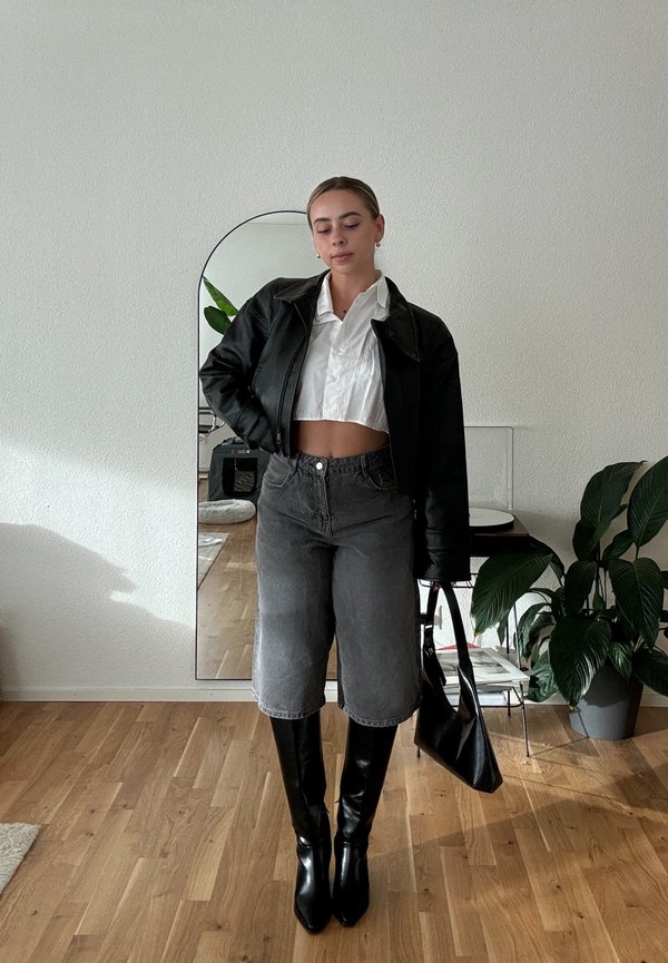 Veste en cuir noire courte, chemise blanche à col, shorts gris taille haute et bottes noires à talons hauteur genou. Sac noir et plantes d'intérieur.