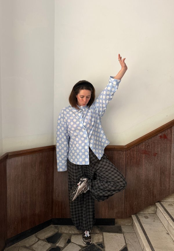 Kvinde i blå prikket skjorte og ternede bukser, der balancerer på ét ben og løfter en arm, stående ved et hjørne af en trappe indendørs.