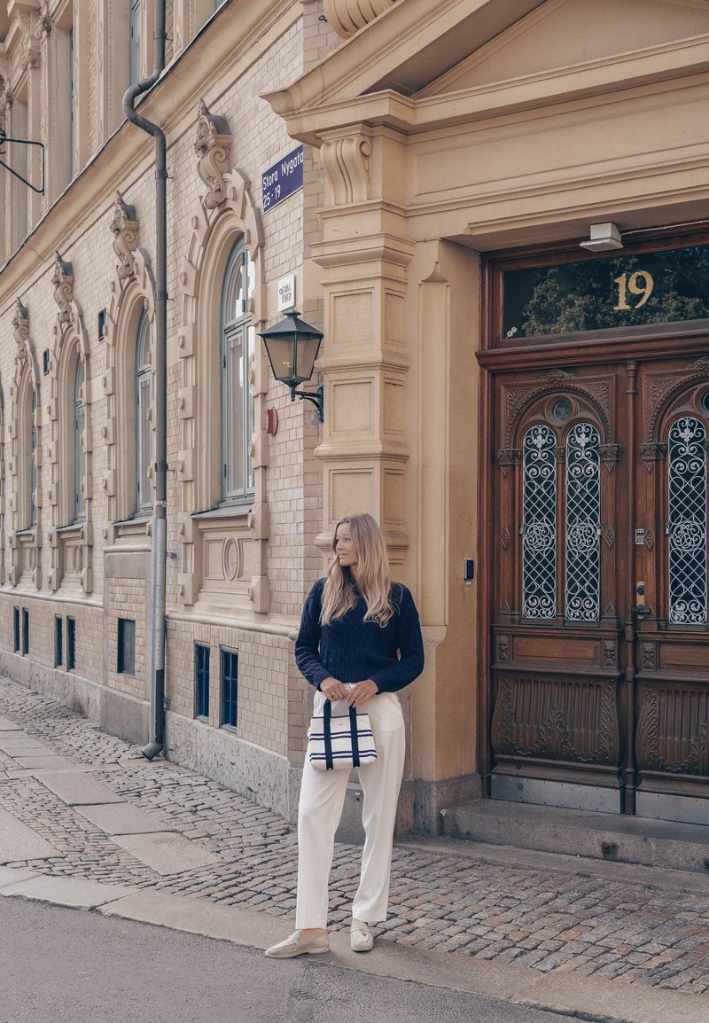 Femme aux longs cheveux blonds, vêtue d'un pull bleu marine et d'un pantalon blanc, tenant un sac rayé, debout près d'une porte en bois ornée et d'un bâtiment historique beige.